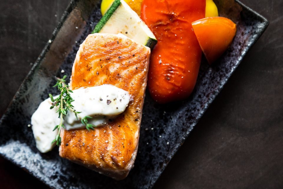 Seared salmon fillet topped with white sauce and thyme, with roasted zucchini, red pepper and yellow tomato on a dark plate