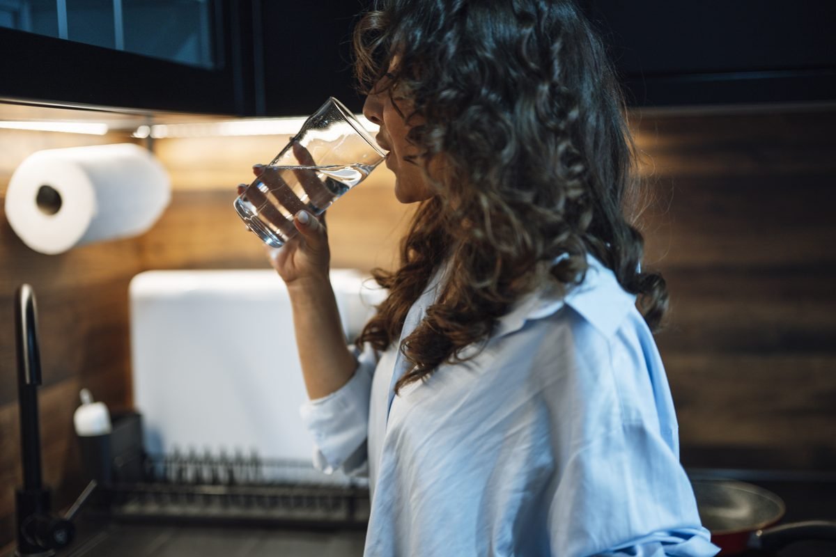 Woman with curly brown hair in a light blue shirt drinking water from a glass in a kitchen.