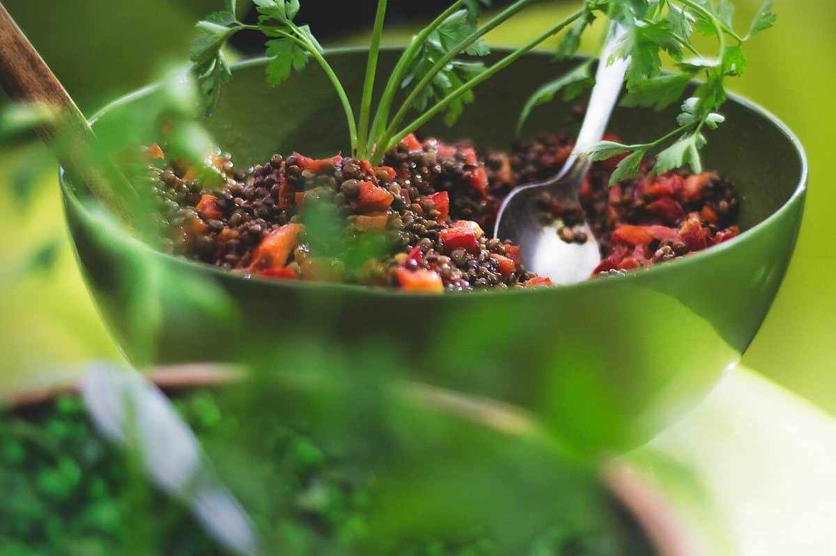 Green bowl of brown lentils mixed with diced red pepper and tomato, garnished with parsley and a silver spoon.