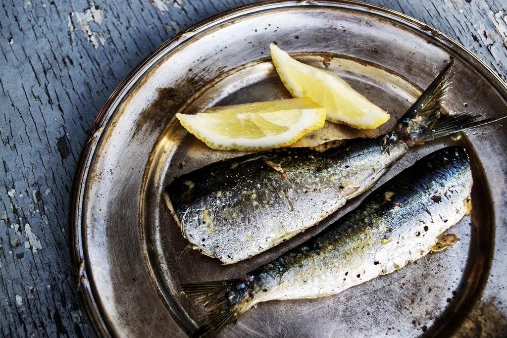 Two whole grilled sardines on a silver plate with two lemon wedges, on a weathered blue wooden table