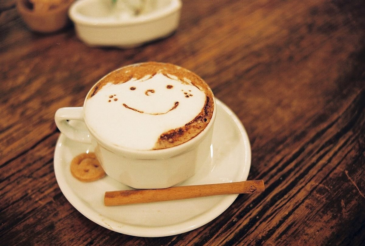 Cappuccino with smiley face foam art on a white saucer beside a cinnamon stick and a biscuit on wood.