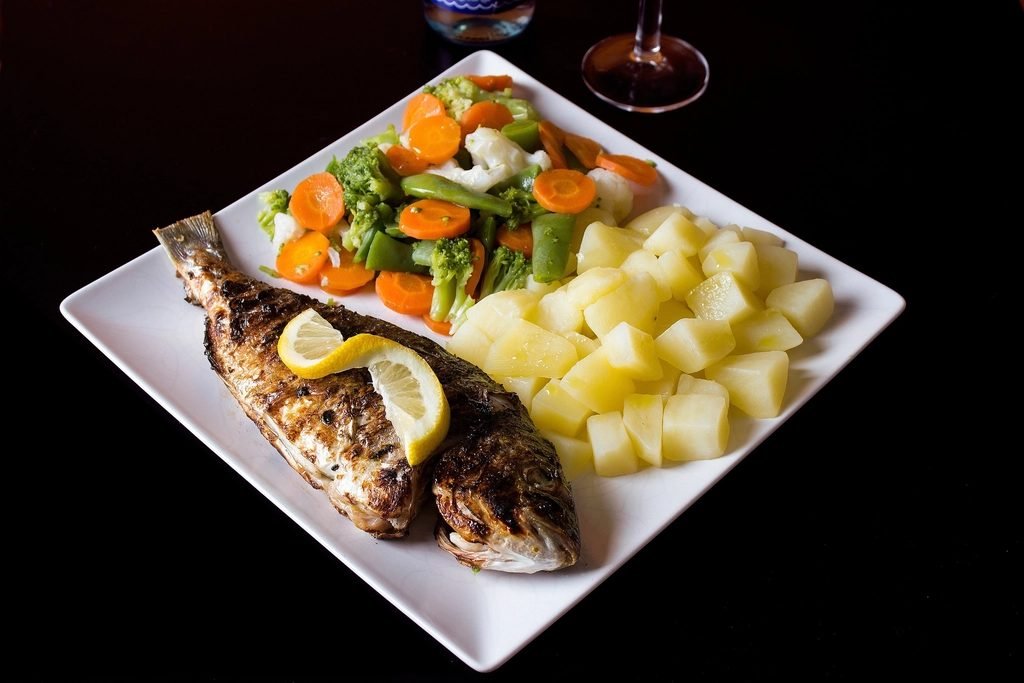 Grilled whole fish with a lemon slice, steamed carrots, broccoli and beans, and potato cubes on a white plate.