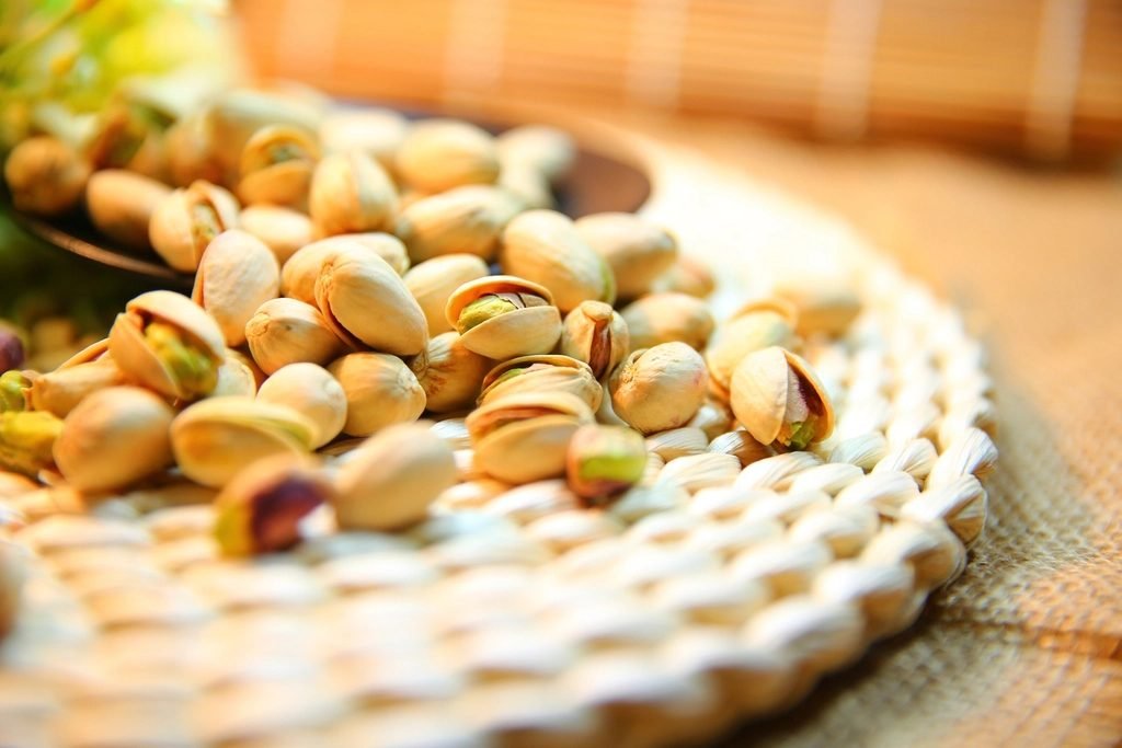 Pistachios in their shells scattered on a round woven wicker mat, with a dark bowl partly visible at the upper left.