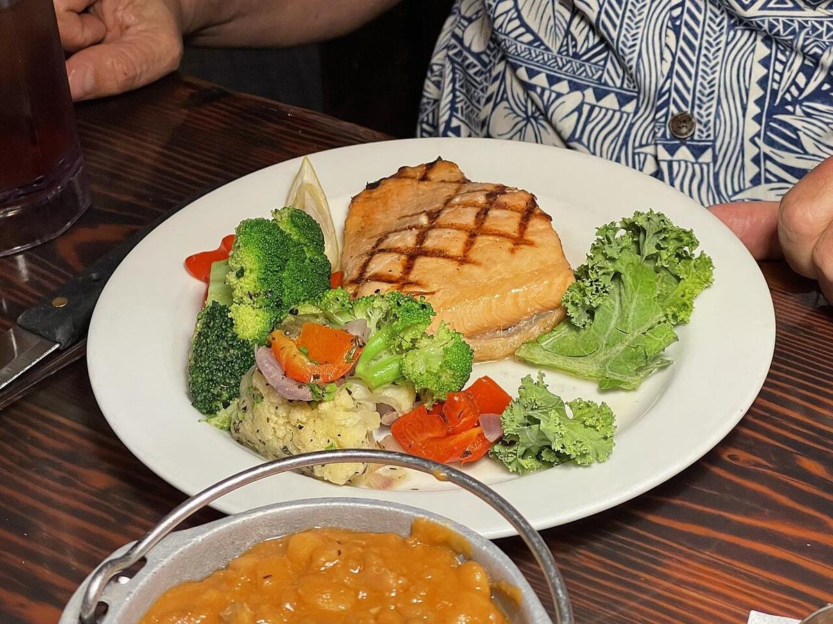 Grilled salmon steak with grill marks beside broccoli, carrots, cauliflower, and kale on a white plate, with a lemon wedge.