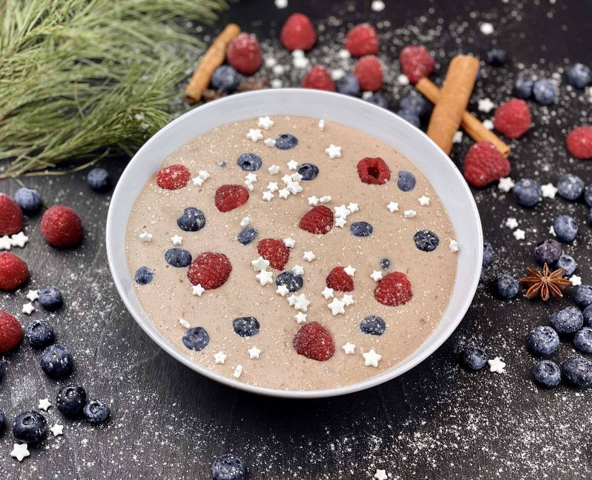 Creamy porridge in a white bowl topped with raspberries, blueberries and star sprinkles, with cinnamon and berries nearby.